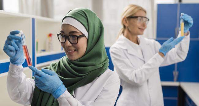 Eine Frau mit einem grünen Schal und Brille hält ein Reagenzglas, im Hintergrund eine zwei Frau mit Schutzbrille und Reagenzglas