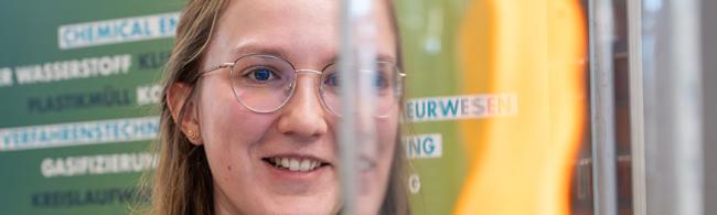Nahaufnahme: Junge Wissenschaftlerin betrachtet eine Flamme in einem Glaszylinder. 