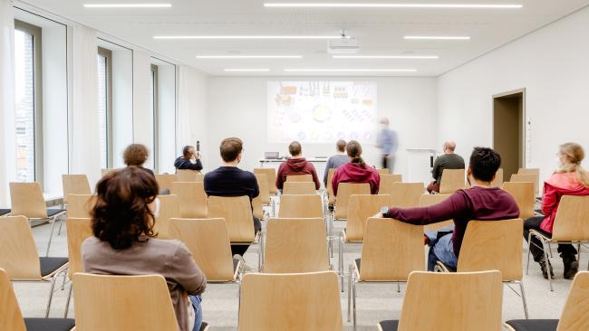 Ein Konferenzraum mit einigen Zuhörer während eines Vortrages. Das Bild ist leicht unscharf